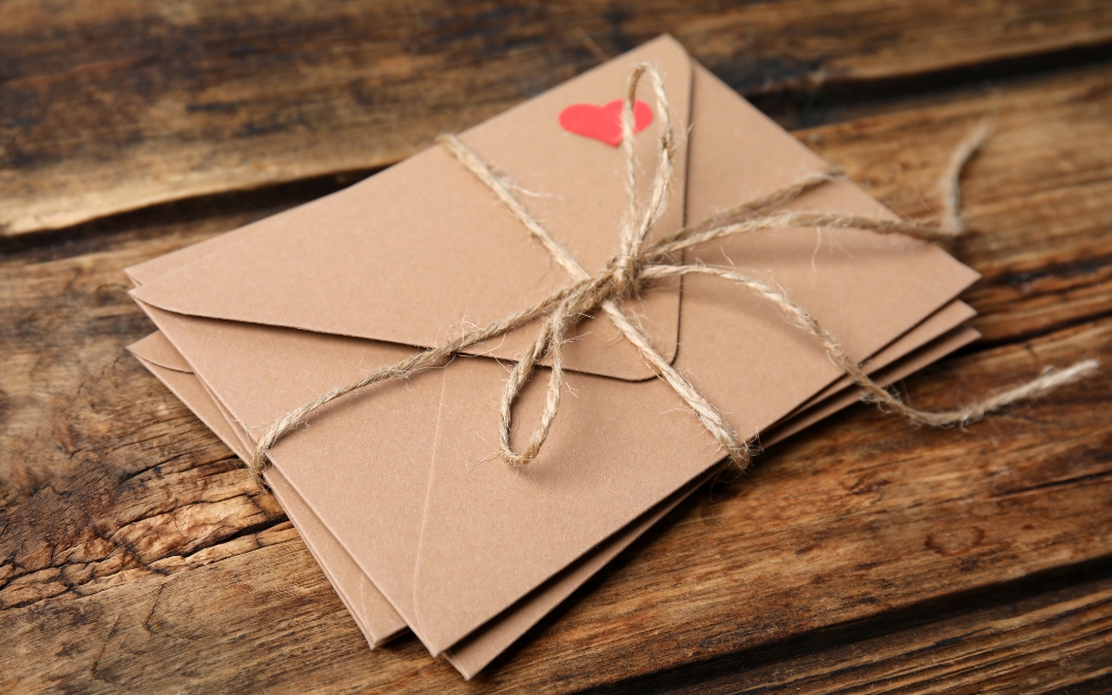 Stack of brown envelopes tied with jute string on a wooden table. Top envelop has a red heart in the right hand corner
