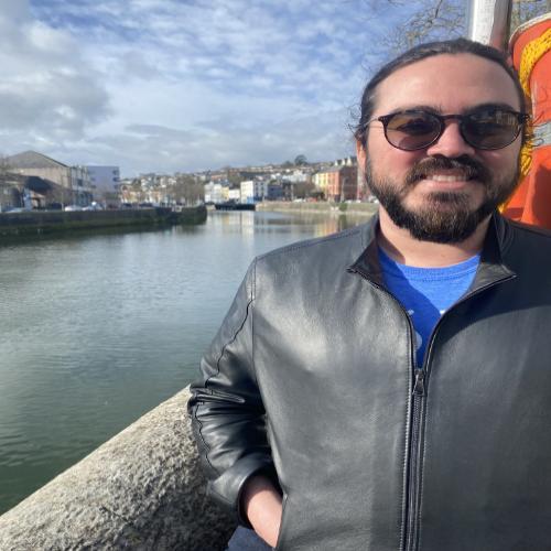 Head and shoulder shot of a smiling white male wearing sunglases and a dark leather jacket zipped up over a blue t-shirt. His dark hair is tied back. Behind him the River Lee stretches out into the urban landscape of Cork City,