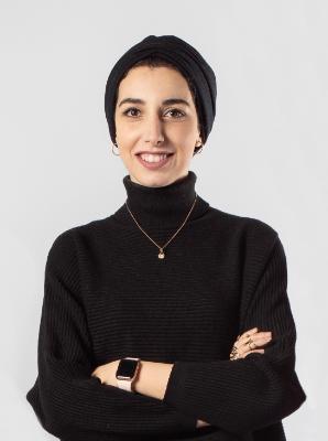 Female student, wearing a black turtle neck jumper and a black headscarf, posing for the camera smiling broadly with her arms folded.