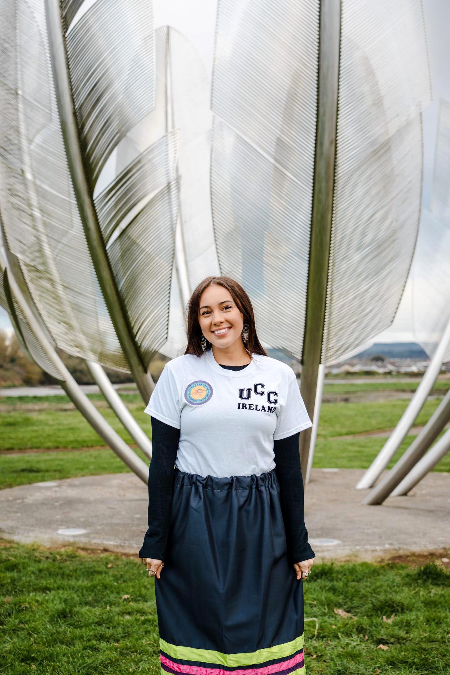 A smiling young female stands in front of a metal sculpture depicting large feathers. She is wearing a white t-shirt emblazoned with the UCC logo and a dark skirt with bright detail at its hem reflecting the Native American Chocktaw Nation.