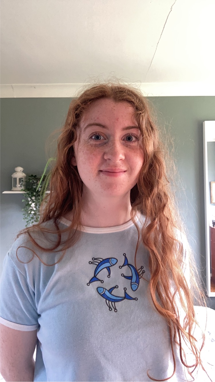 Student wearing a light teeshirt decorated with three fish, and with long red wavy hair, smiles for the camera.