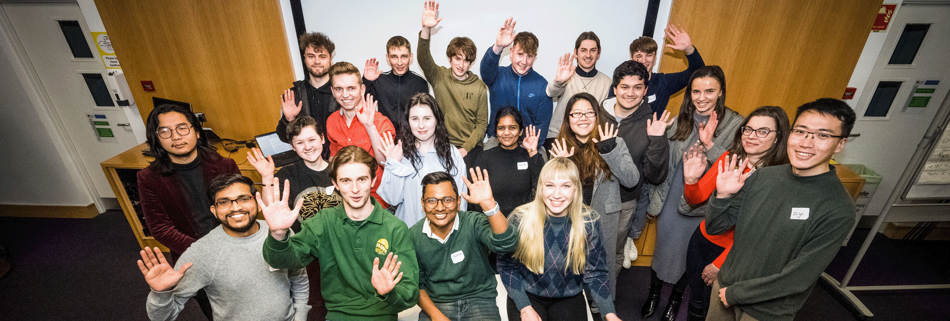 High angle photo of group of young people in a room waving towards camera
