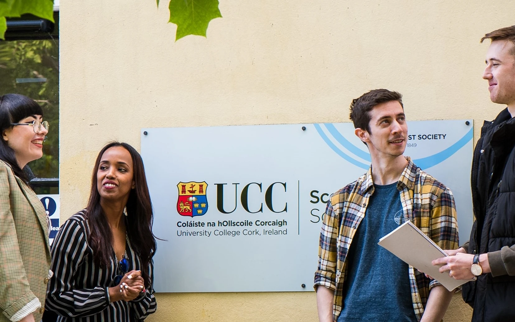 Four students chatting in front of a wall-mounted sign featuring the UCC crest