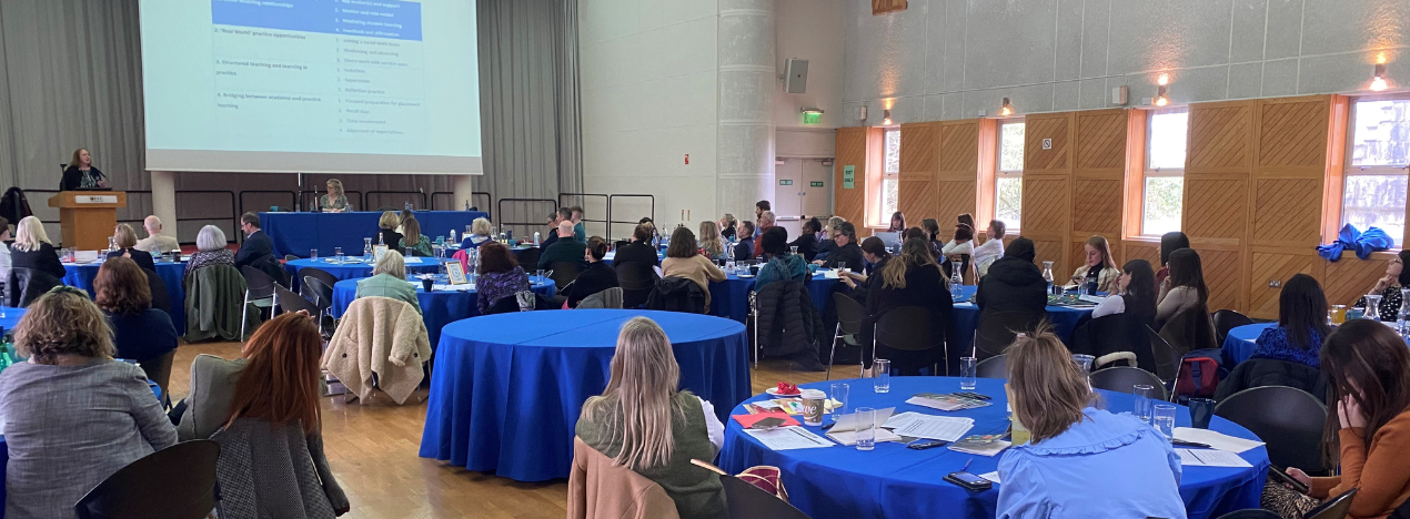 Image of large room with tables full of people watching speakers at an academic conference