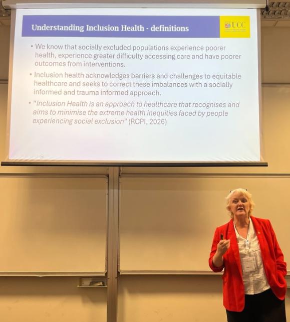 Dr Flynn stands at the front of a lecture hall, speaking beneath a projected slide titled “Understanding Inclusion Health - definitions.” The slide explains how socially excluded populations experience poorer health outcomes and describes inclusion health as an approach that seeks to reduce health inequities through equitable, socially informed, and trauma-informed care. A University College Cork (UCC) logo appears in the top-right corner of the slide. The presenter wears a bright red jacket and a conference badge and gestures while addressing the audience.