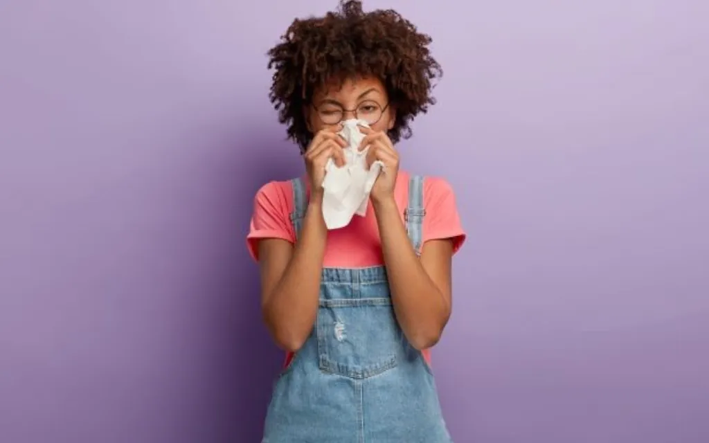 Person with curly hair and round glasses holding a tissue to their nose against a light purple background. The person is wearing a pink top with denim straps and is positioned in the centre of the image, with ample empty space around them suitable for banner text.