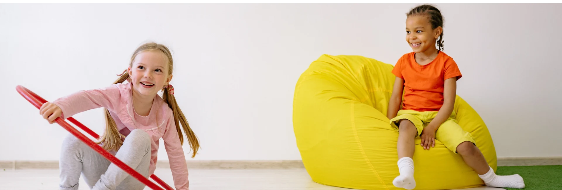 Two children in a bright indoor room. One child in a pink long-sleeve top and grey trousers sits on the floor holding a red hula hoop, while another child in an orange T-shirt and yellow trousers sits on a large yellow beanbag chair. The room has a light-coloured floor, a white wall, and ample open space.