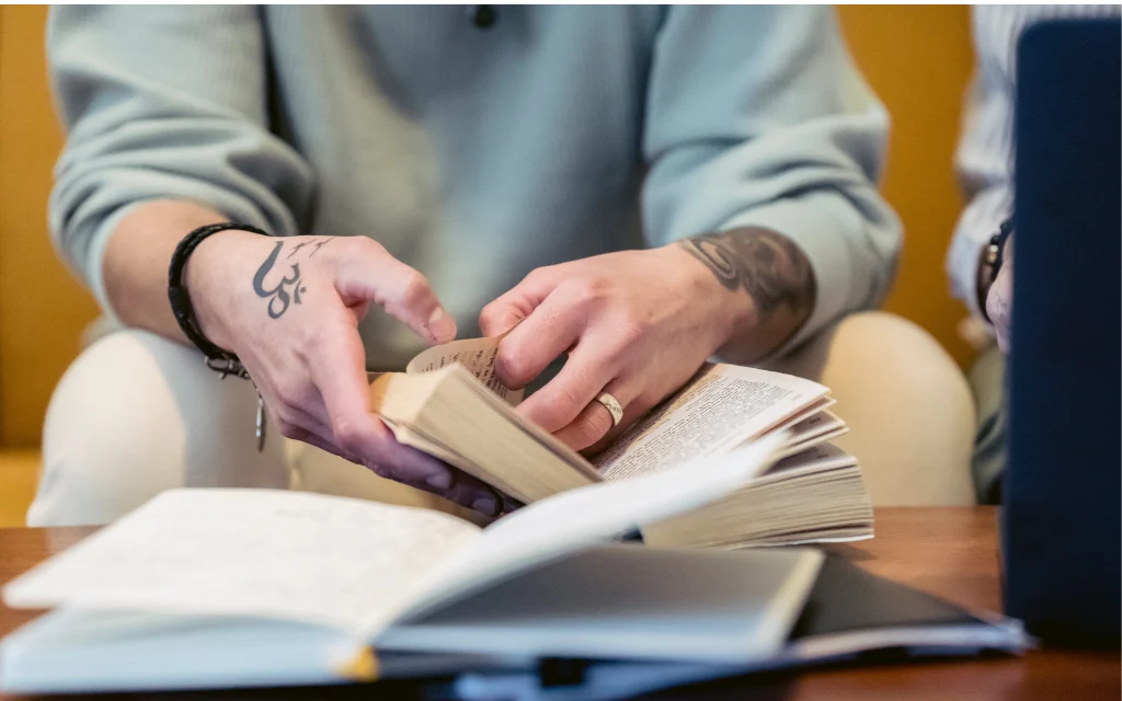 Close-up of a person’s tattooed hands turning the pages of an open book while seated. The image focuses on the book and hands, with a shallow depth of field creating a softly blurred background. A bracelet and ring are visible, and the scene is lit with warm indoor lighting.