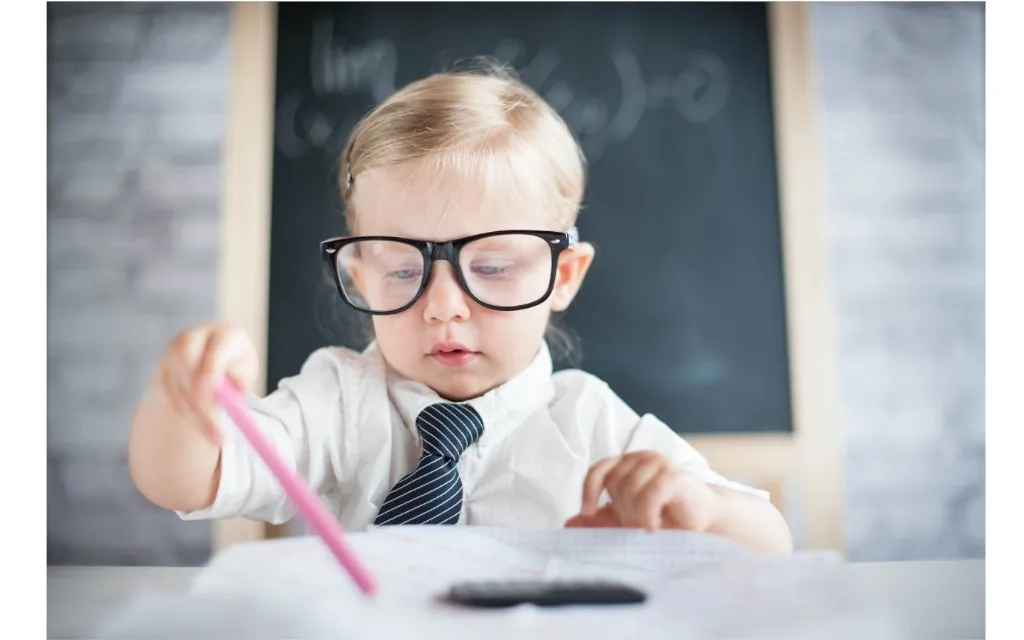 Young child wearing oversized black glasses, a white shirt and striped tie, concentrating on writing with a pink pencil at a desk. A chalkboard is visible in the softly blurred background, creating a classroom or learning-themed setting.
