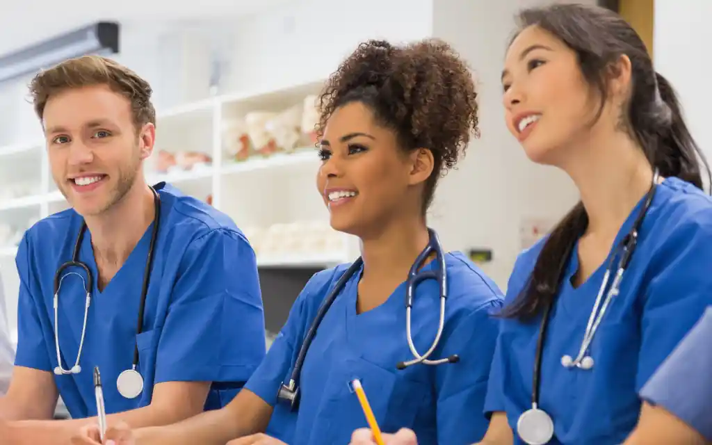 Four healthcare students wearing blue scrubs and stethoscopes sit together in bright clincial classroom.  One person wears a white coat over scrubs.  Shelving and teaching materials are visible in the background.  The group appear engaged in a disucssion facing toward the same area of the room