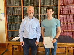 Student with cert standing next to lecturer (book backdrop)