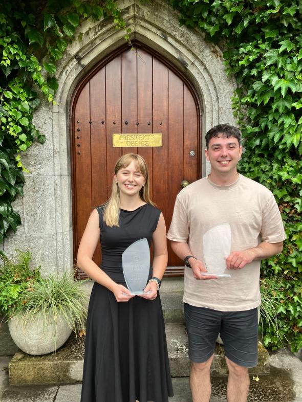 NUSOD Award Winners, two students holding certs in front of wooden door with green foilage