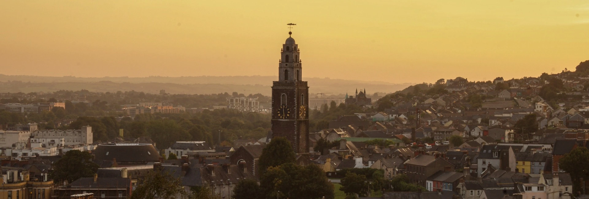 Panoramic view of Cork City at sunset, featuring the clock tower of St. Anne’s Church rising above densely packed rooftops. Warm golden light fills the sky and casts a soft glow over the historic cityscape, with rolling hills, residential neighborhoods, and distant buildings fading into a hazy horizon.