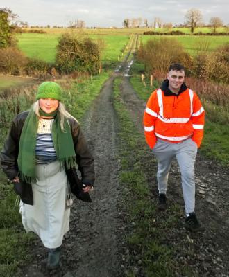 Two people dressed warmly and smiling, walking on a muddy track towards the camera. Green landscape with watery sunshine behind them.