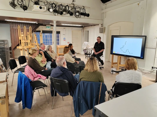 A group of people listen to a person speak at the top of a room A group of people listen to a person speak at the top of a room