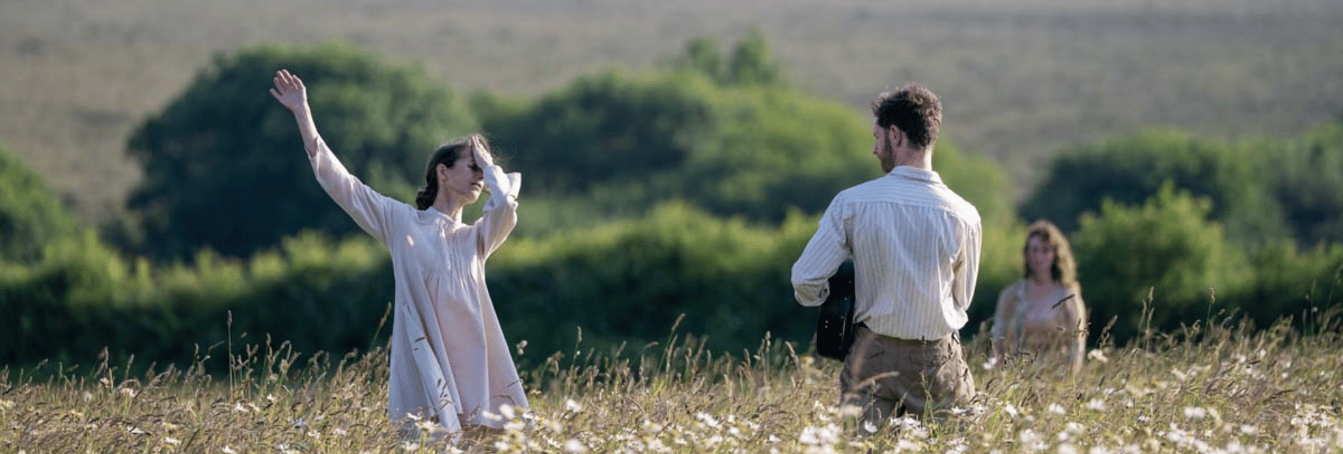 Three people stand in a field of wildflowers. One person in the foreground on the left has their arm raised. Another person in the middle is holding a guitar and facing away. A third person is in the background on the right.
