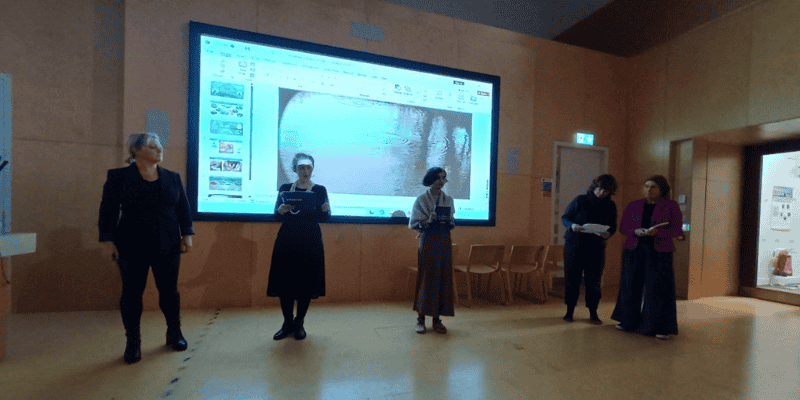 A group of women stand in front of a screen in The Shtepps. The room is dark and lit from the screen