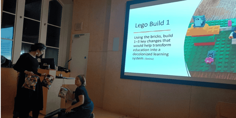 Two women hold bags of Lego bricks in front of a screen describing a Lego build in The Shtepps