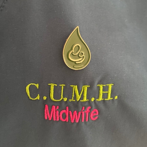 Close up of a metal pin the shape of a teardrop on scrubs . Placed over text 'CUMH midwife'