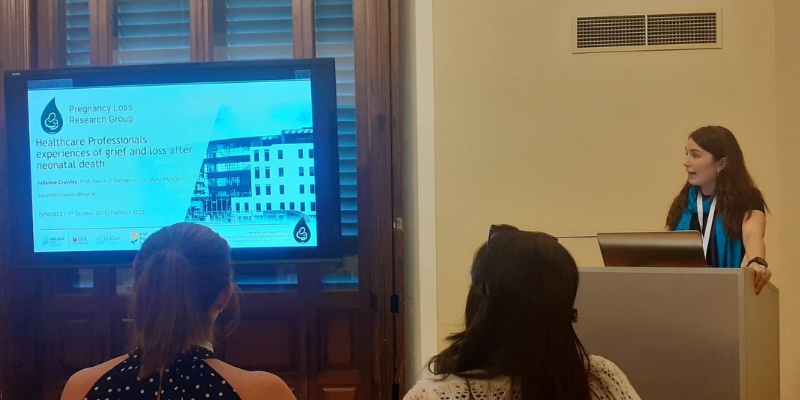 Women speaking behind a podium with a large presentation screen to the left of the photograph