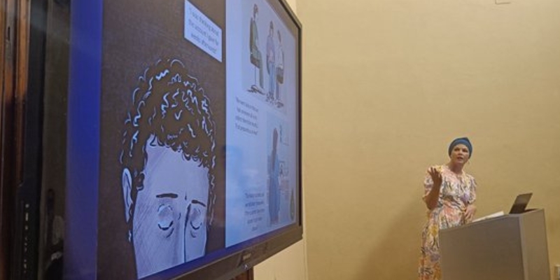 Woman speaking behind a podium with a large screen to the left of the photograph