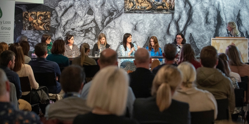 Six people sitting in a row at a table as part of a panel discussion, with one person standing at a podium chairing the discussion