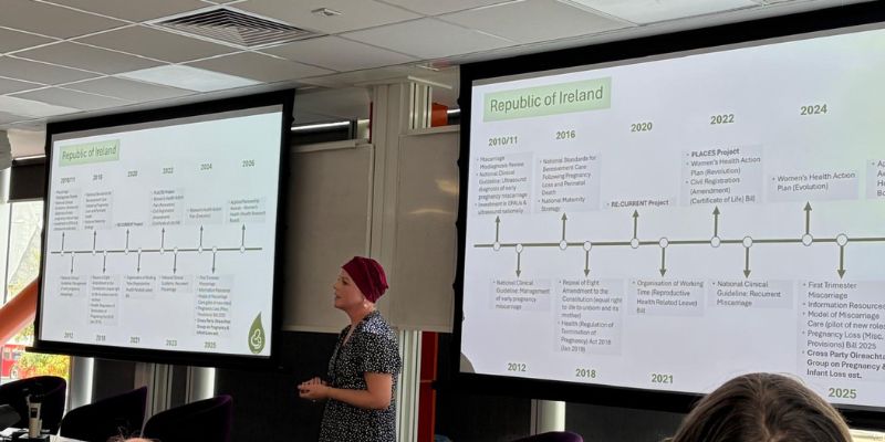 Woman (standing) speaking at a conference, two large presentation screens behind her
