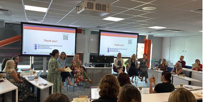 Panel discussion at a conference - woman (chair) standing, five women seated on high chairs in fromt of two large presentation screens; audience visible around them Panel discussion at a conference - woman (chair) standing, five women seated on high chairs in fromt of two large presentation screens; audience visible around them