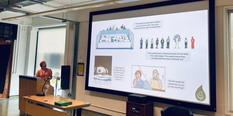 Woman standing behind a podium with a large presentation screen to her left, with images from 'Why my baby died' on it