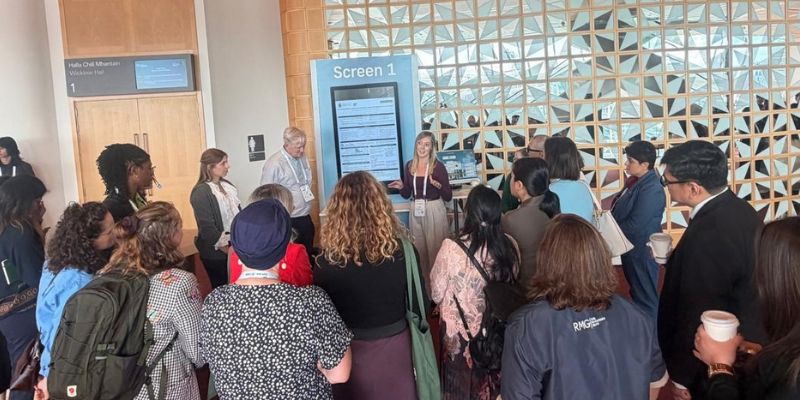 Group of people standing around a mobile presentation screen listening to a woman speaking to the crowd Group of people standing around a mobile presentation screen listening to a woman speaking to the crowd