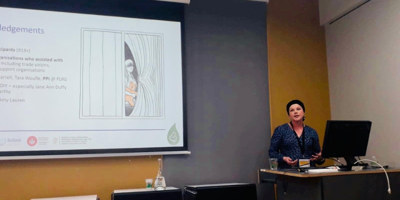 woman speaking at a conference with large screen to the left