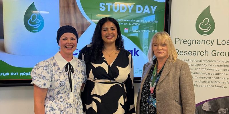 Three women standing, smiling, in front of a large presentations screen that says 'Study Day' with Pregnancy Loss Research Group pull-up banner in the background