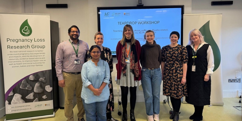 Seven people standing smiling in front of a large presentation screen with a large pull-up banner that says 'Pregnancy Loss Research Group' to the left of the photo