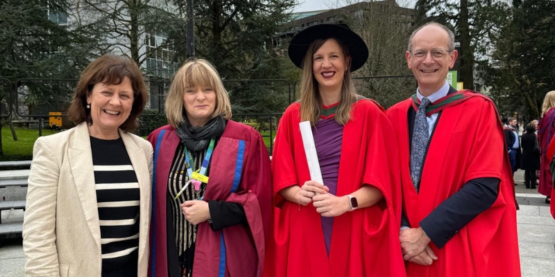 Four people standing for a photograph on graduation day; three in red robes