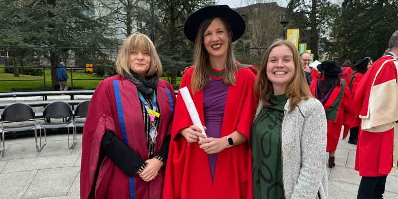 Three people standing for a photograph on graduation day; two in red robes