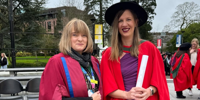 Two people standing for a photograph on graduation day; both in red robes, one holding a scroll