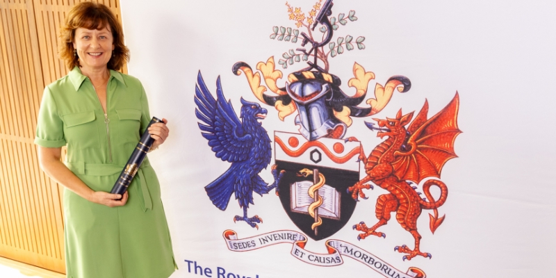Woman in a green dress holding a scroll beside a large white banner with a large coloured logo of the Royal College of Pathologists on it