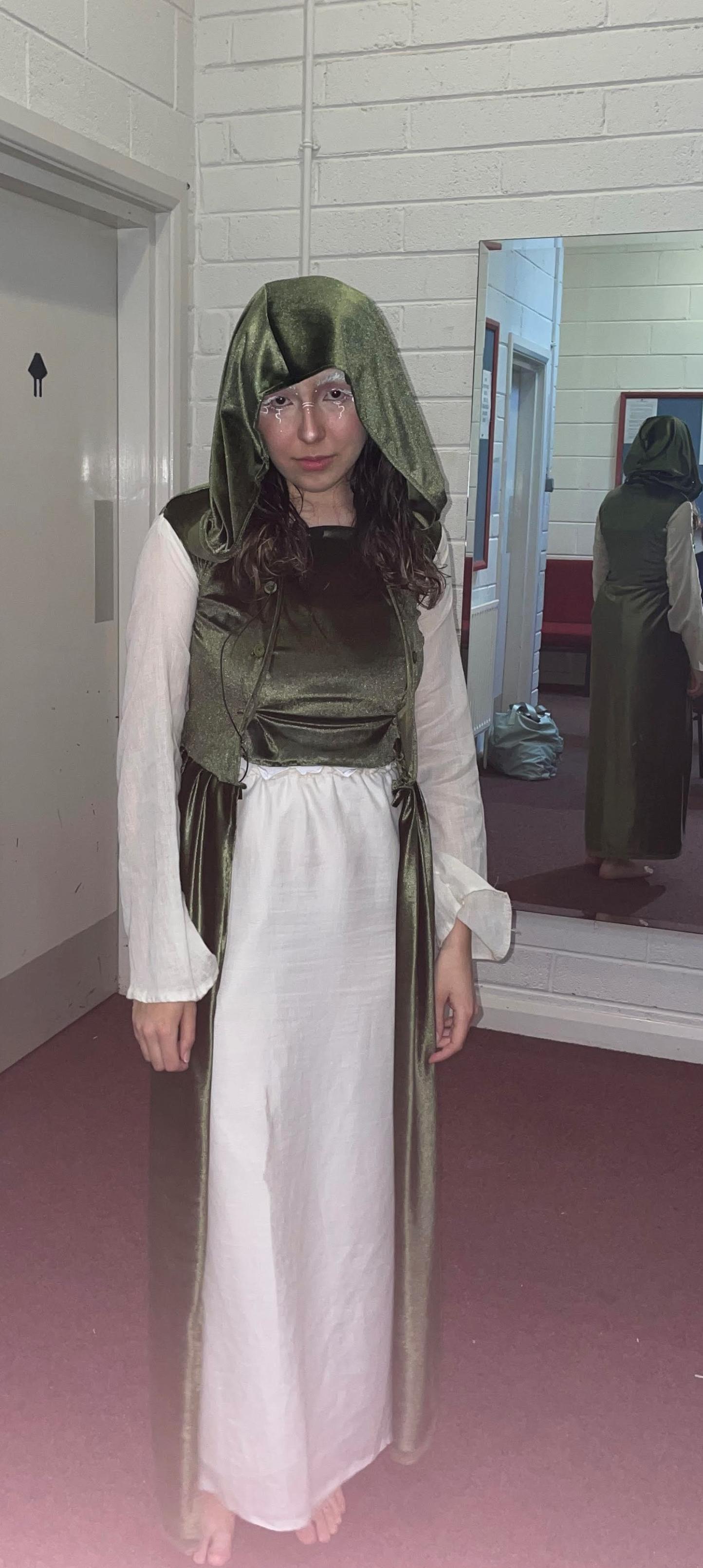 Student in green and white costume with hood. Long brown hair frames her face as she stares at the camera.