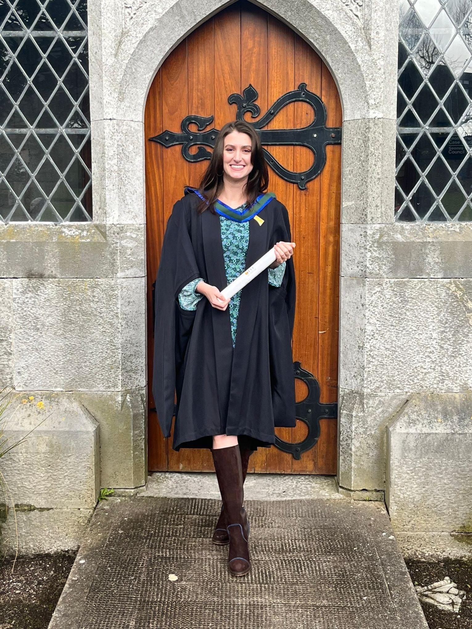 Smiling student with long dark hair, wearing a black gown over a blue dress, and holding a parchment scroll, stands on stone steps in front of an arched wooden door.