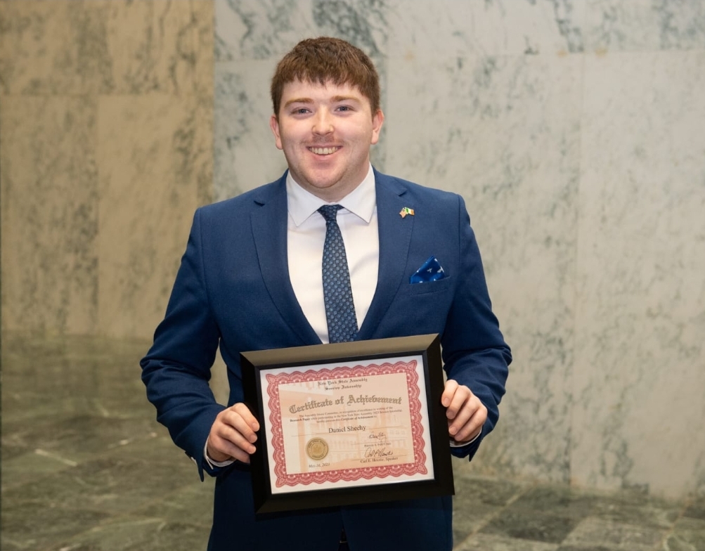 Smiling student in a dark suit holding up a certificate for the camera