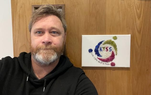 Head shot of male student with grey-flecked beard and wearing a black hoodie leaning against a brown door facing the camer.