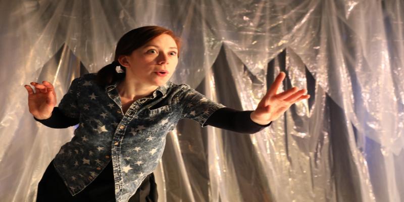 Young lady with long red hair in a blue shirt decorated with white stars against a plastic backdrop. She is reaching forward towards the viewer with a compelling look of storytelling on her face.