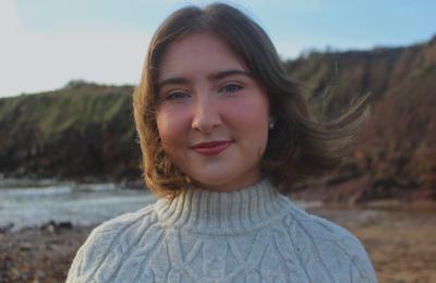 Headshot of young girl with short brown hair wearing a light grey jumper. She is shown smiling for the camera against the backdrop of a sea scape.