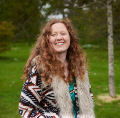 Female with long curly red hair, wearing a colourful cardigan, standing in a grassy area with trees, smiling directly at the camera.