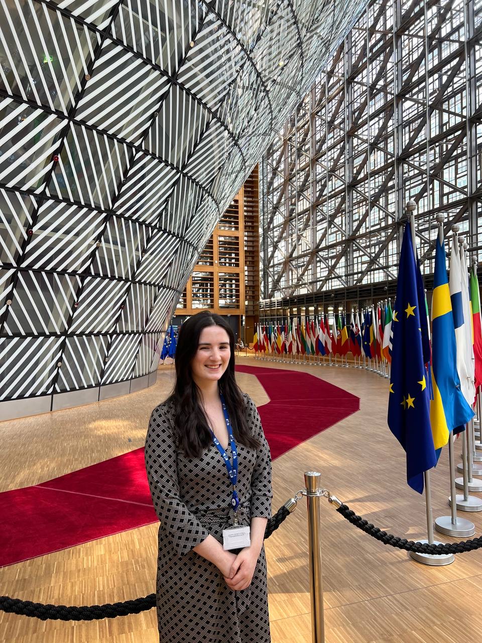 Woman with long dark hair standing in front of the large steel-structured internal facade of an office building. The corridor is lined with European flags.