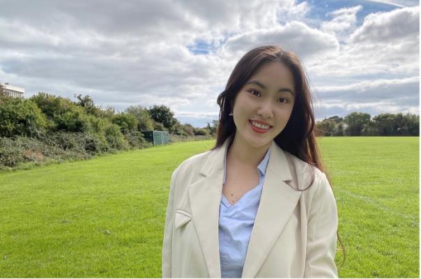 MA Teaching Chinese to Speakers of Other Languages female student, in a cream coat and light blue top, smiling for the camera against the backdrop of a green field.