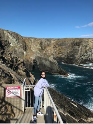 MA Teaching Chinese to Speakers of Other Languages female student, in sunglasses, light blue sweatshirt and denim jeans, smiling for the camera against a rugged coastline backdrop.