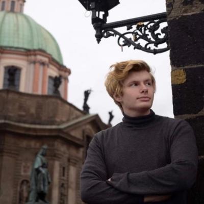 White man with red hair, wearing a dark roll-necked jumper, is standing by a wall starting off into the distance.