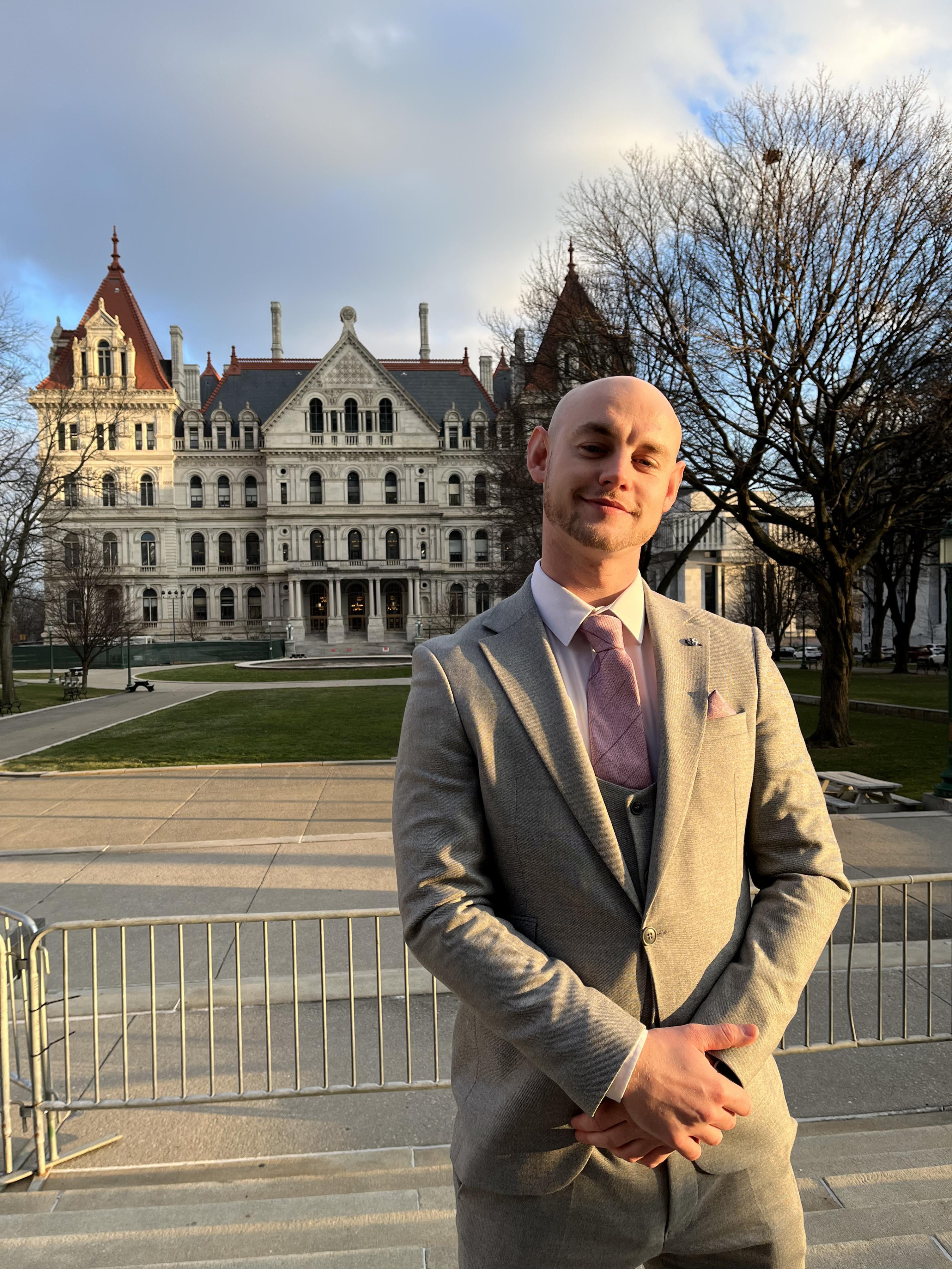 Young man in a light-coloured suit poses in front of an ornate building.