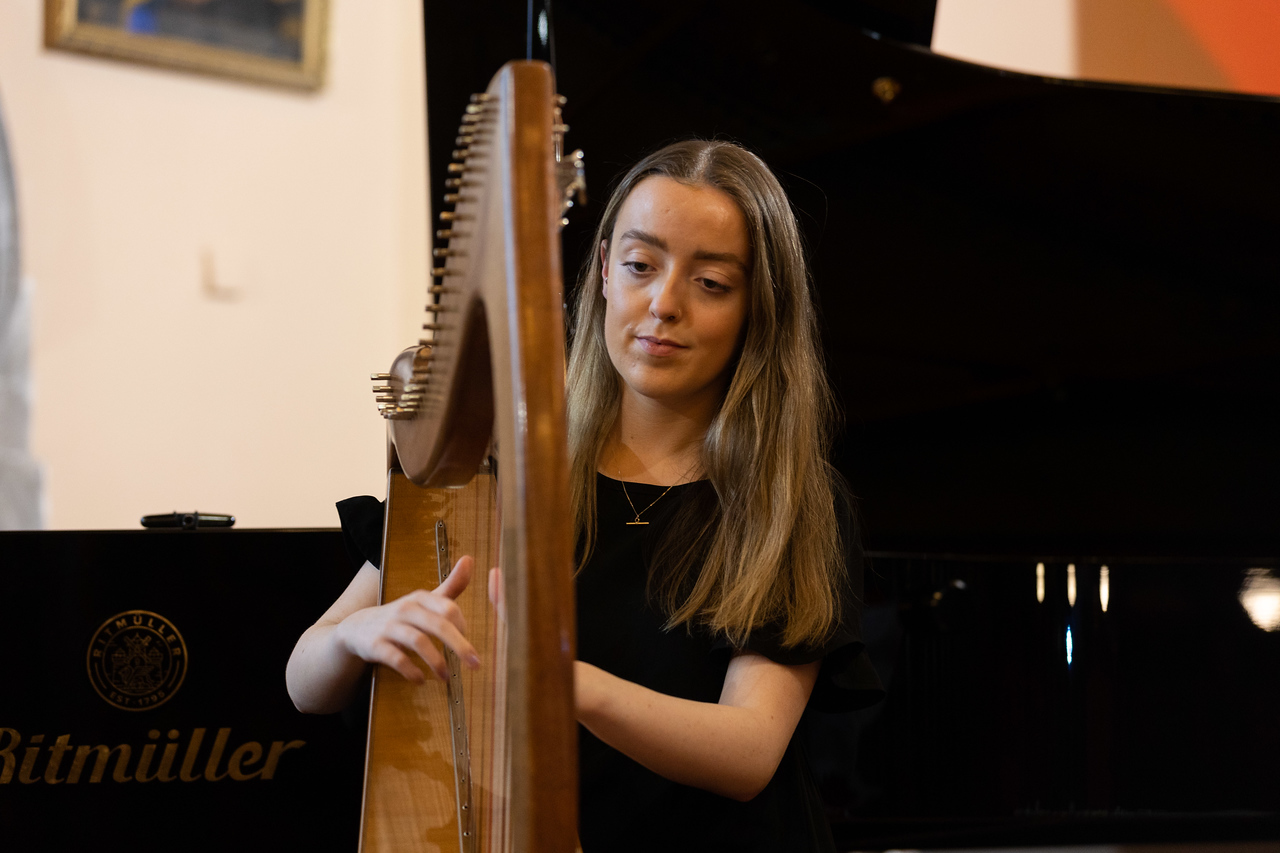 Young woman with long brown hair in a dark dress, plucking on the strings of  a brown wooden harp.